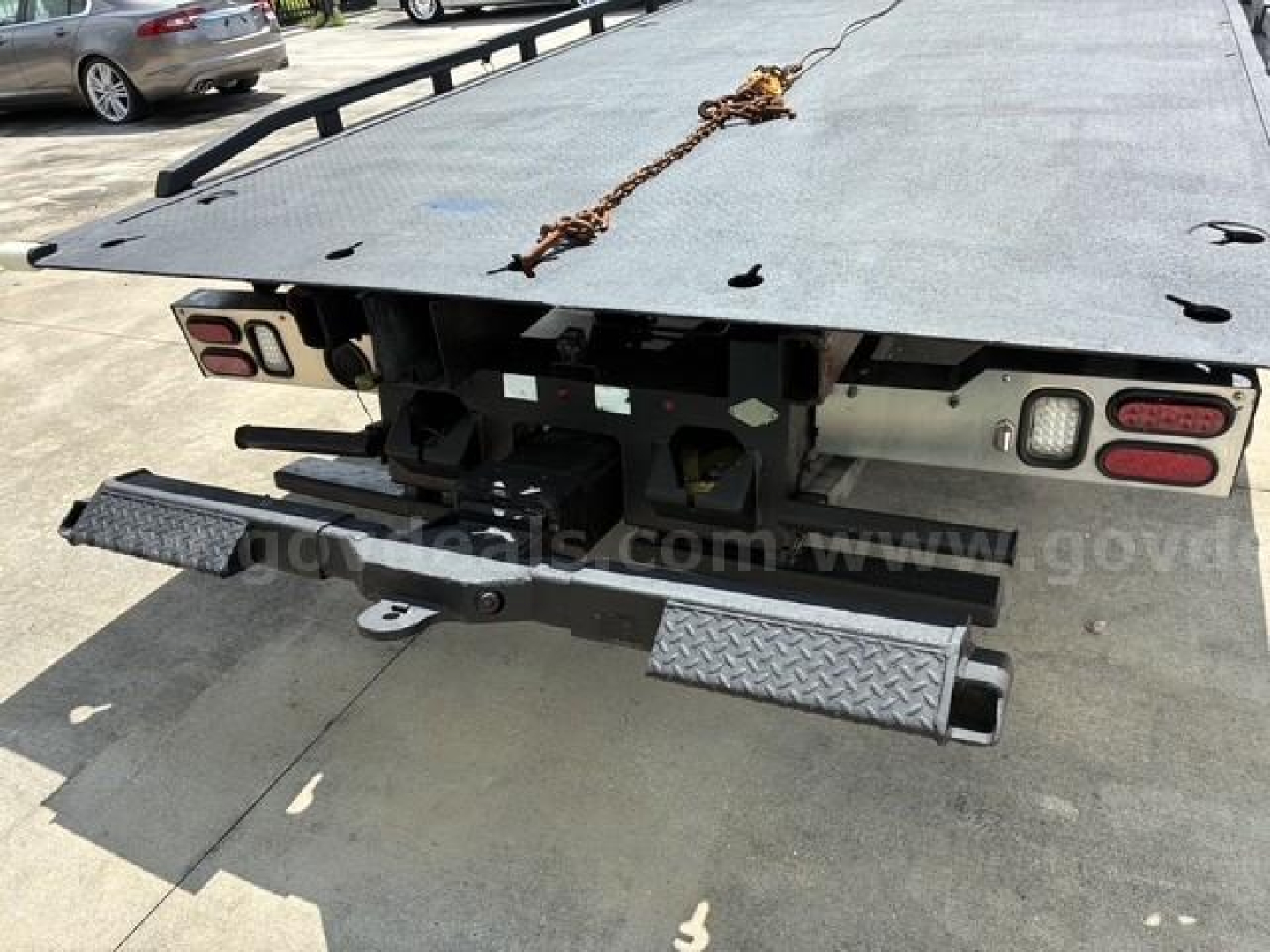Rear view of a 2017 Freightliner M2 106 flatbed truck showing bumper, taillights, and a diamond plate step; preliminary text visible on the tailboard and a chain attached.