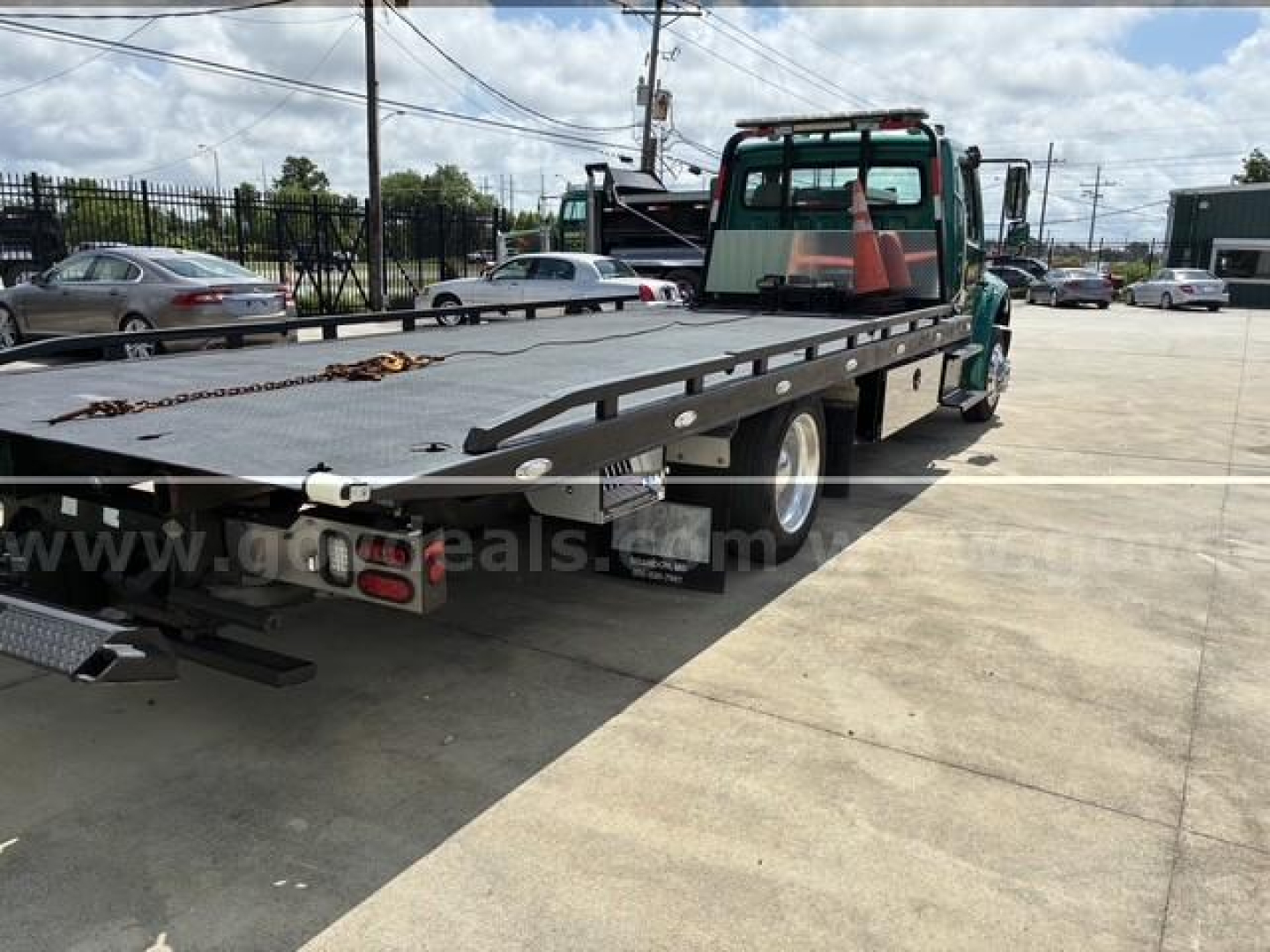 Flatbed tow truck with a green cab and long black steel bed; visible markings include a chain on the bed and rear lights; signage or decals on the flatbed are partially obscured.