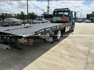 Flatbed tow truck with a green cab and long black steel bed; visible markings include a chain on the bed and rear lights; signage or decals on the flatbed are partially obscured.