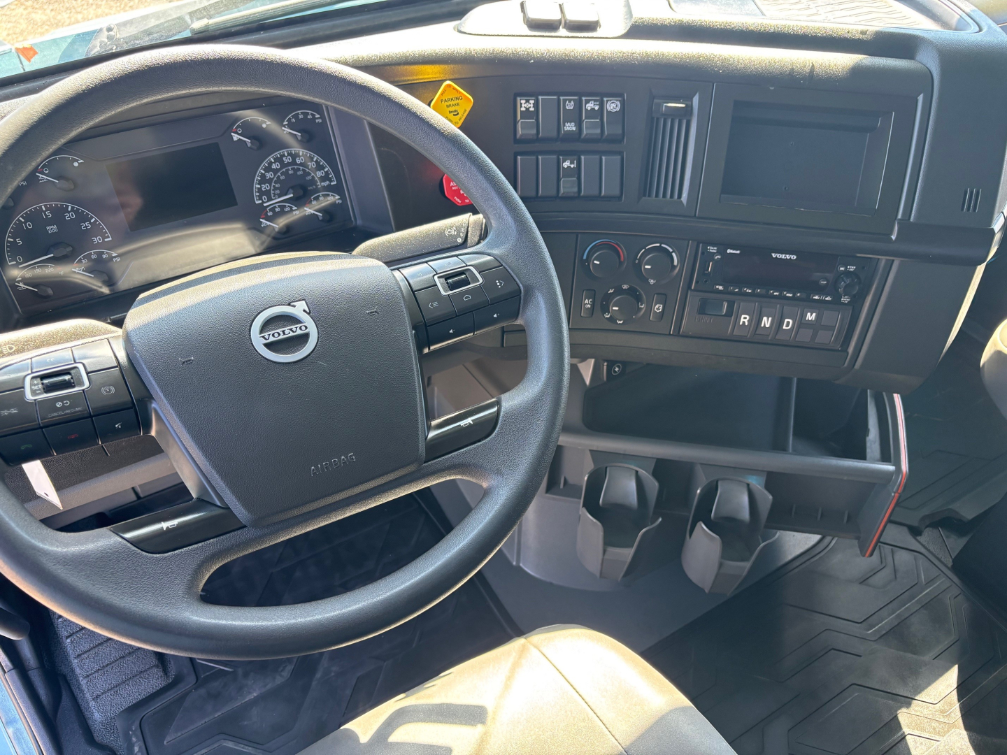 Volvo truck interior showing steering wheel with Volvo logo, dashboard, gauges, gear selector, and center console; text not visible clearly.