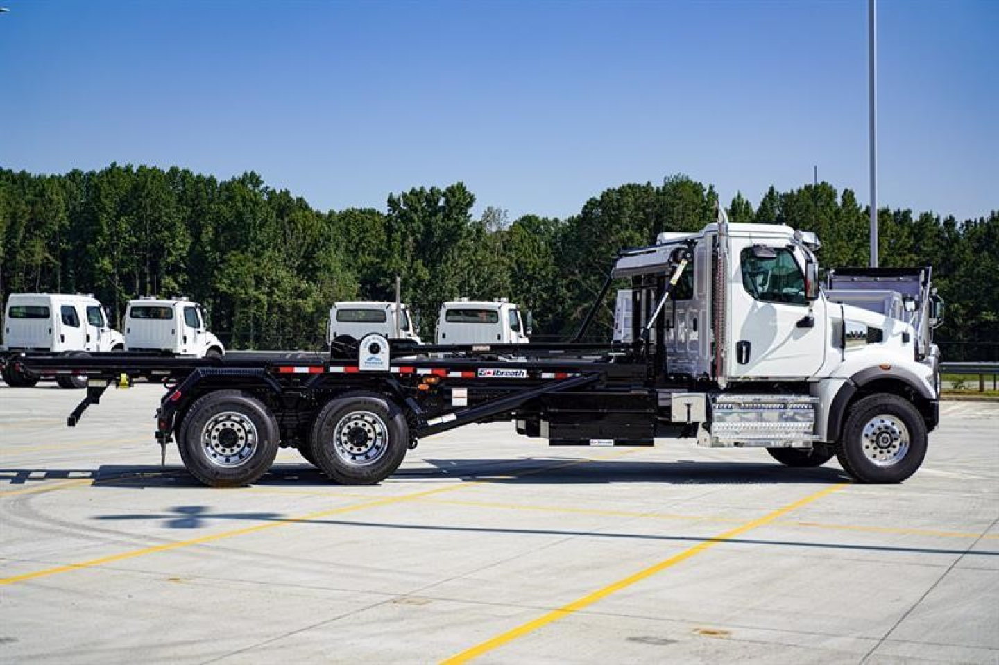 Western Star 47X roll-off truck in white with long chassis and dual rear axles, parked on a lot with trees in background.