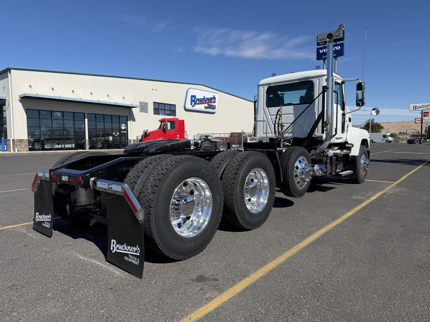 2027 Mack PI84T tractor with Manhattan-style sleeper, three rear axles, chrome wheels, and Bracken Trucks mud flaps; dealership building visible in background.