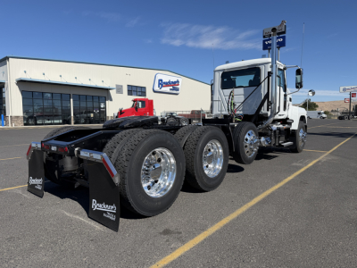 2027 Mack PI84T tractor with Manhattan-style sleeper, three rear axles, chrome wheels, and Bracken Trucks mud flaps; dealership building visible in background.