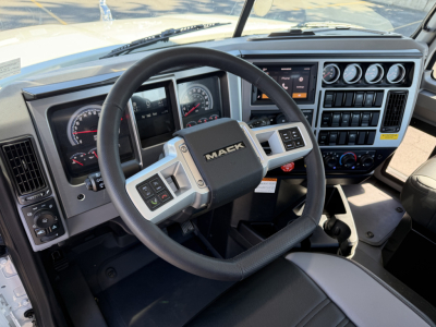 Truck cockpit with Mack steering wheel and dashboard gauges; controls and digital display visible.