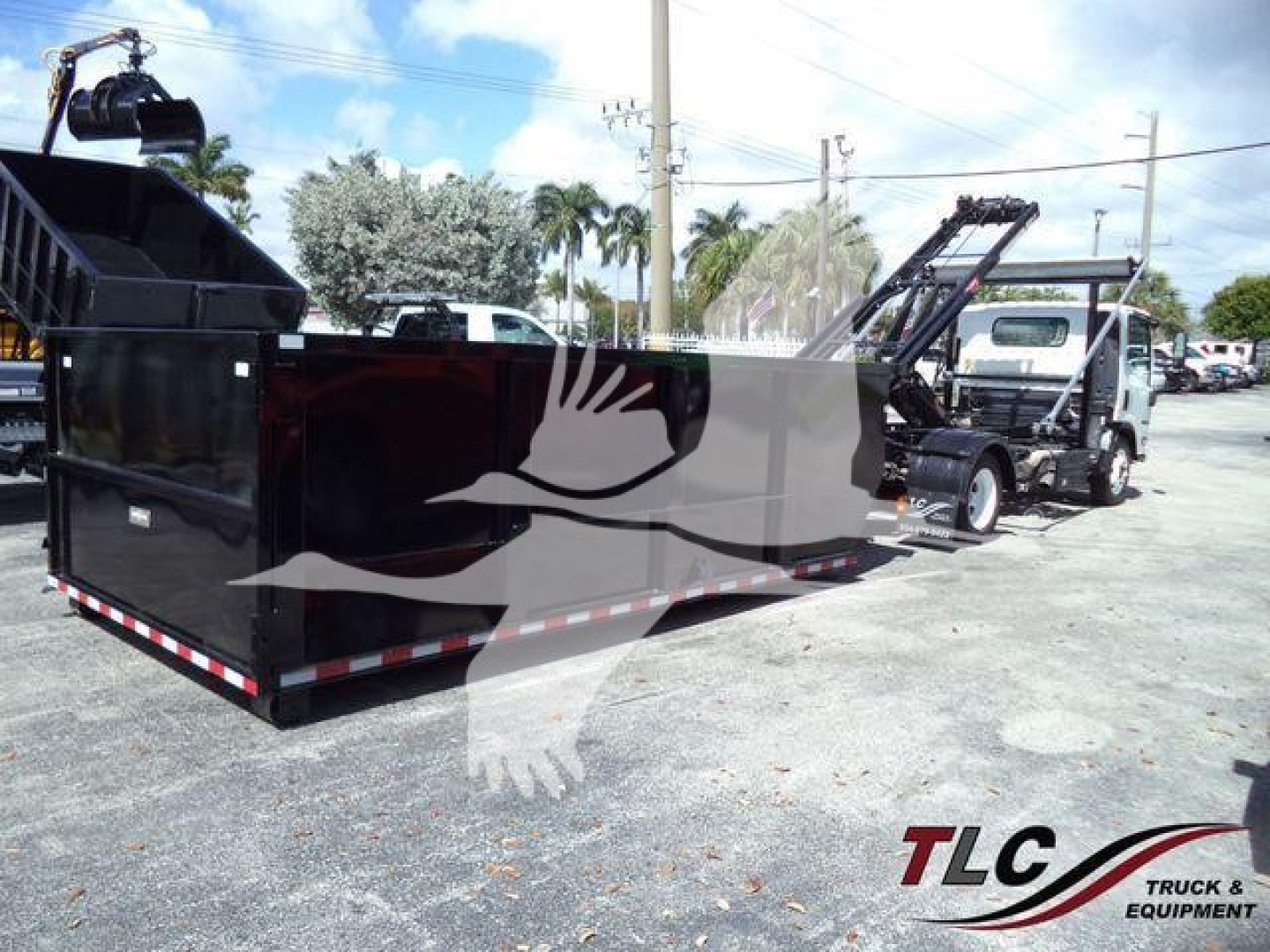Isuzu NPRR roll-off truck with large black outer container and loading hoist in yard; visible TLC Truck & Equipment logo watermark.