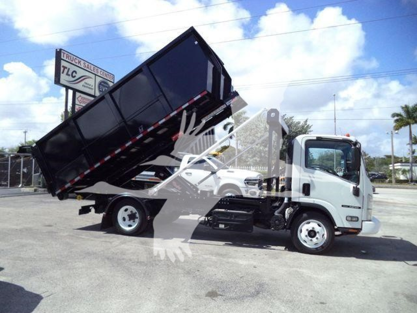 Isuzu NPR medium-duty truck with a raised black dump bed in a tipping position; side view shows cab front with Isuzu badge, yard site with sign in background; no visible decals or serials readable