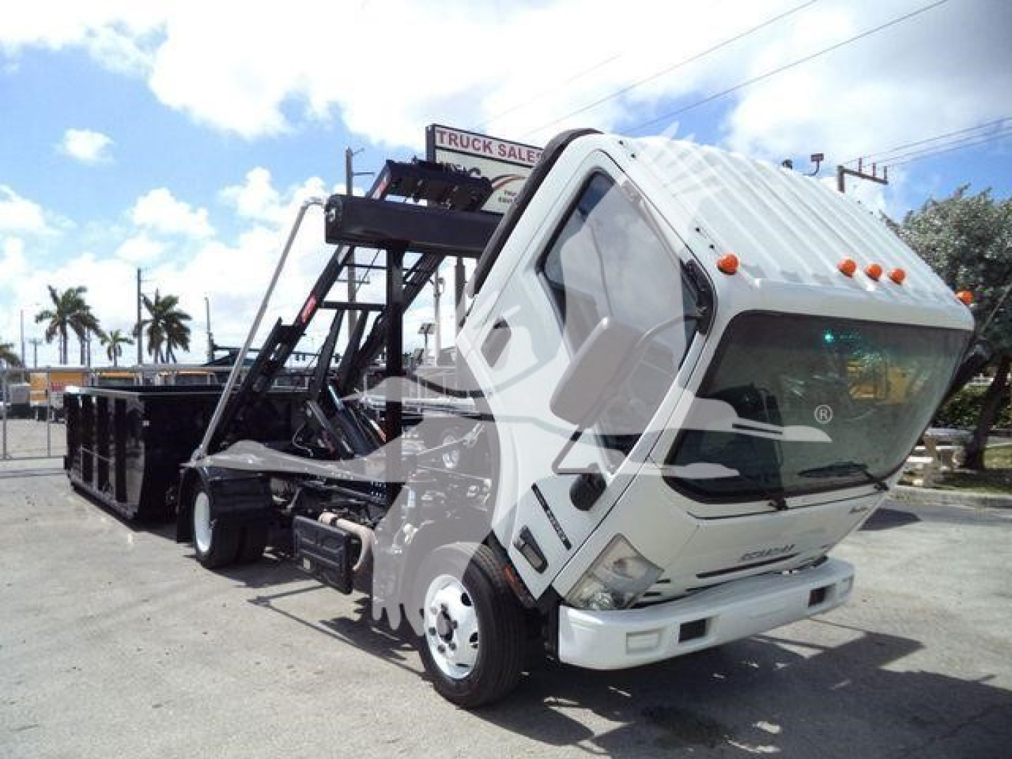 Isuzu NPR/RR truck body in tilt cab configuration in a lot. Front bumper visible with windshield, side mirrors, and hydraulic arms for roll-off bed. Some signage in background.