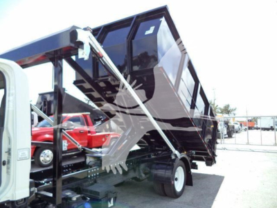 Isuzu NRR roll-off truck tail with hydraulic dump bed raised, showing a white cab and black dump body in a yard.