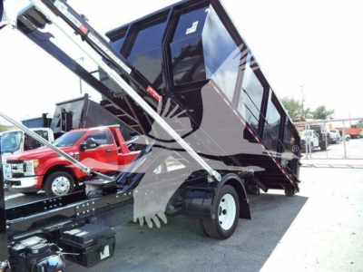 Isuzu truck in a roll-off configuration showing a tipped black cargo container on a truck bed; hydraulic arm and winch visible; surrounding vehicles in lot.