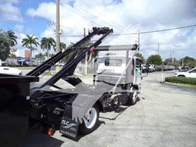 Isuzu NPR flatbed tow truck with a hydraulic hooklift arm in upper position, white cab, black chassis, side view showing rear wheels and equipment, storage boxes, and safety lights.