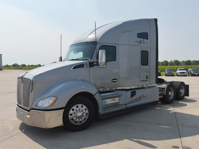 Kenworth T680 semi-truck cab and chassis in a parking lot, silver exterior with sleeper cab, side steps, and dual rear axles.