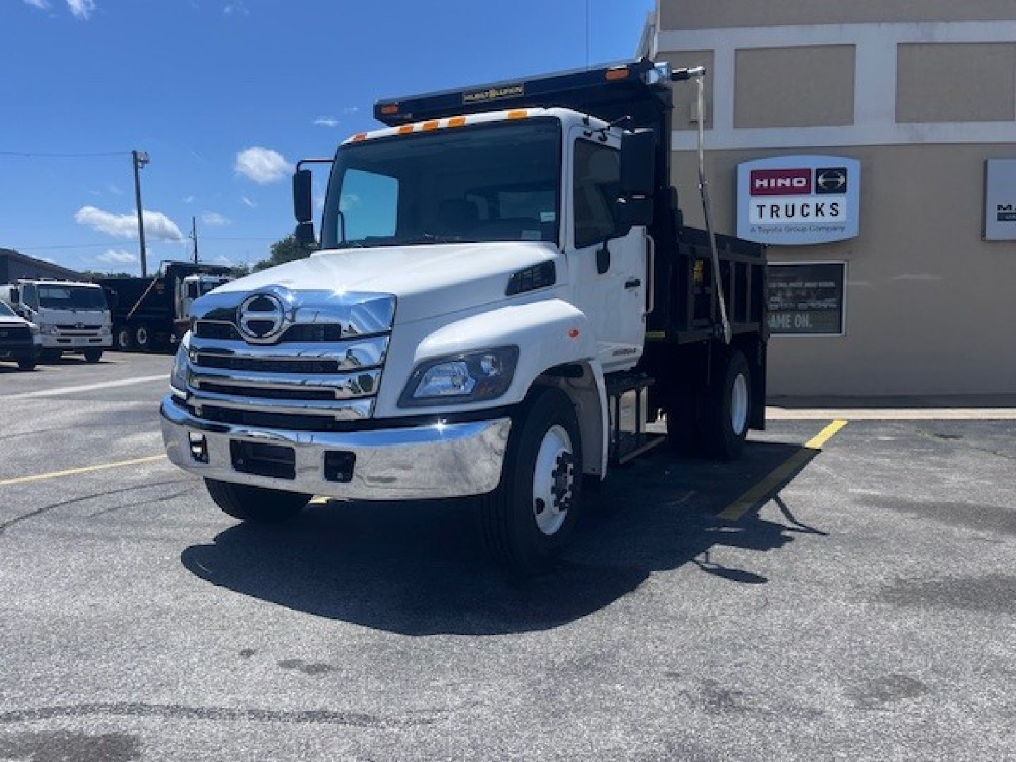 White 2026 Hino L7 dump truck in a dealership lot, front three-quarter view showing chrome grille, Hino emblem, and dump bed, with sign reading HINO TRUCKS in background.