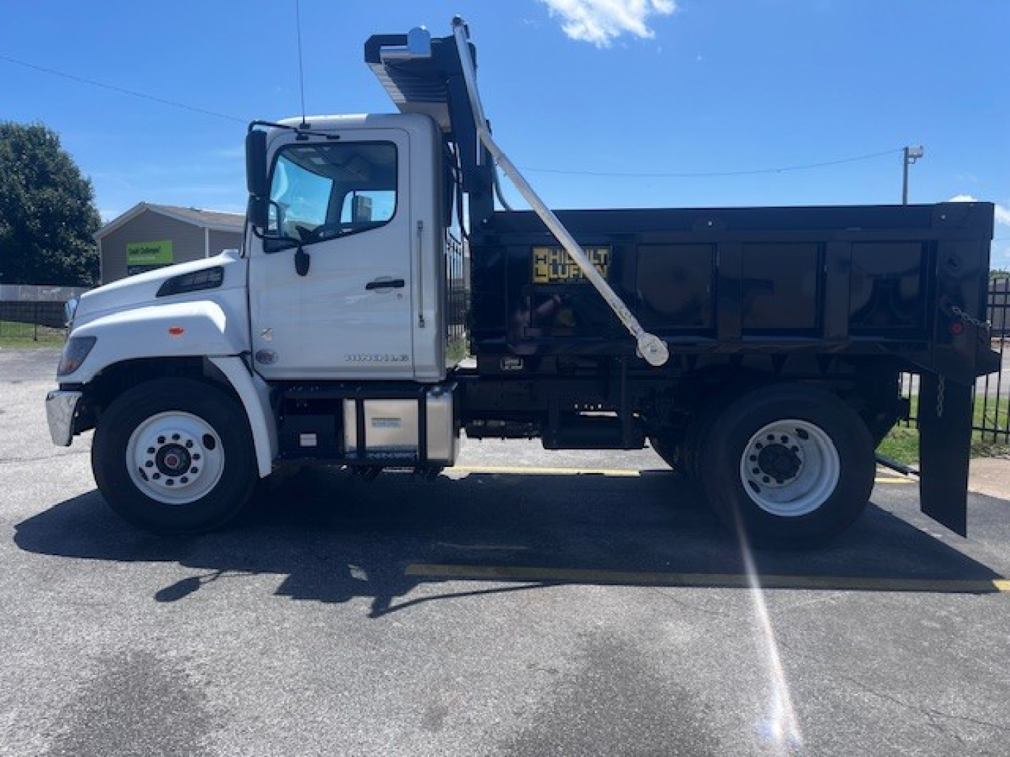 Dump truck in side profile with white cab and black dump bed, Hino L7, medium-duty, with a mounted hydraulic crane arm over the bed.