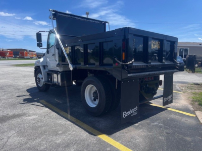 Dump truck shown from rear-left side with black dump bed, white cab, and large mud flaps reading Bueschman? branding; truck has chain tether and hydraulic bed action.