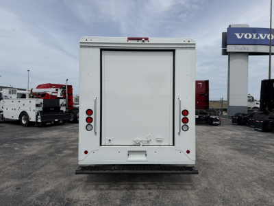 Rear view of a white step van with a large cargo compartment. There are two vertical taillights on each side, a rear bumper step, and a rear door with handles. The truck is in a dealership lot with other commercial vehicles in the backgroun