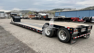 Trailer showing a long lowboy flatbed with dual rear axles and green cabover in background; side markings and reflection; visible dealership lot.