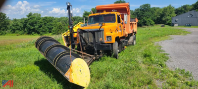 Orange dump truck with a large yellow plow/wing attachment in a grassy lot. Cab over, plow blade in front, black coil spring suspension, trees and a gray building in background.