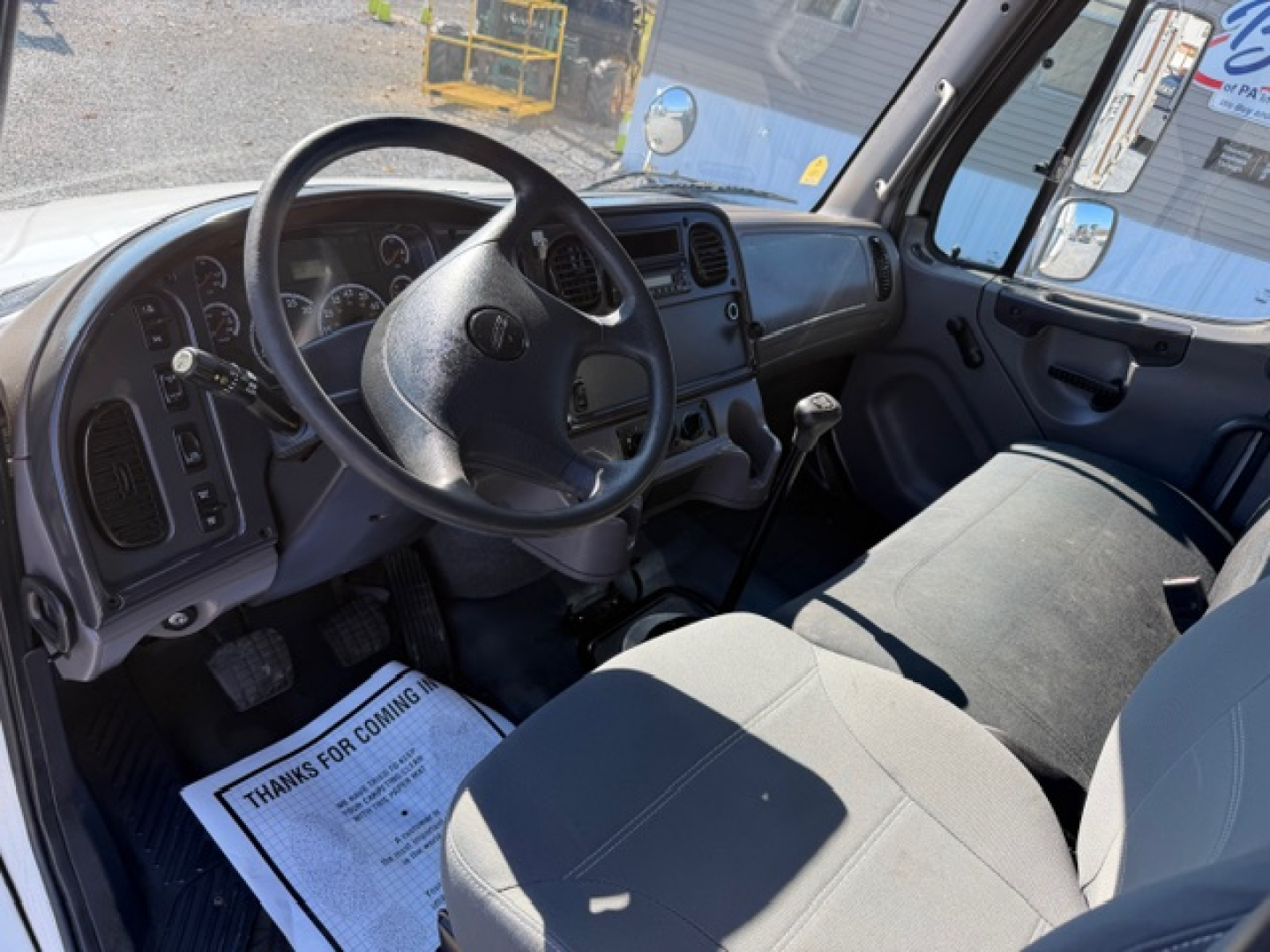 Cab interior of a 2009 Freightliner M2 106 showing steering wheel, manual gear shifter, and driver and passenger seats. Dashboard with gauges and center console visible.