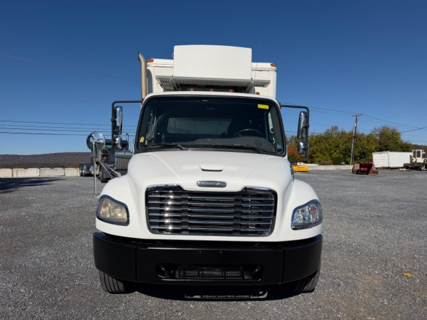 2009 Freightliner M2 106 truck front view. White cab with large grille, refrigerator unit on top, black bumper. Side mirrors extend out. Clear sky.