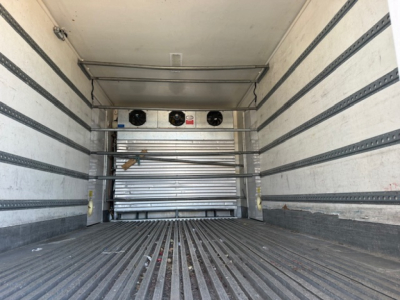 Refrigerated truck cargo area showing empty trailer with metal side walls and ribbed floor. At the far end, a refrigerated unit with three circular fans and a metal door.