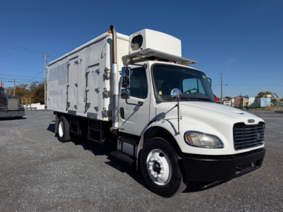 Freightliner M2 106 refrigerated truck left-front view showing cab and cargo box with white exterior and refrigeration unit on top.