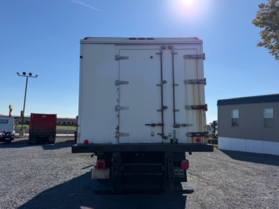 Back of a white refrigerated Freightliner M2 106 box truck showing rear doors with multiple latch hinges and bumper, light posts and a building in the background.