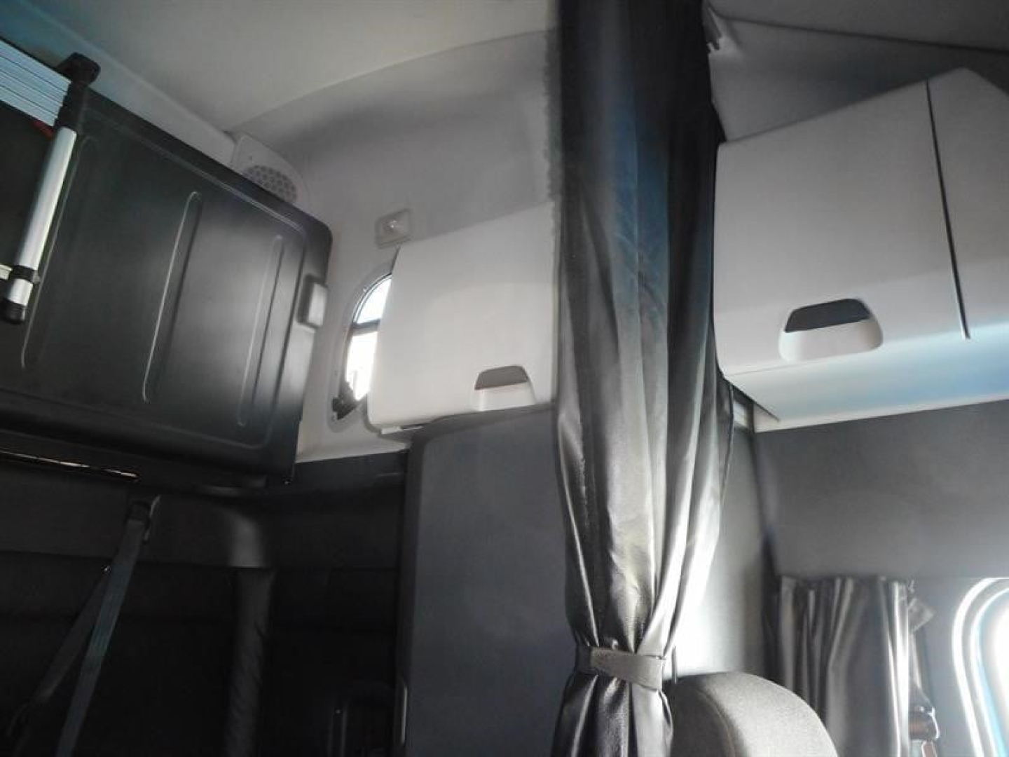 Cab interior of a Freightliner Cascadia sleeper showing upper storage cabinets and a sun visor/partition, with curtains and a window