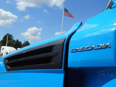 Front corner of a blue 2023 Freightliner Cascadia 126 truck showing grille and Cascadia badge on the side.