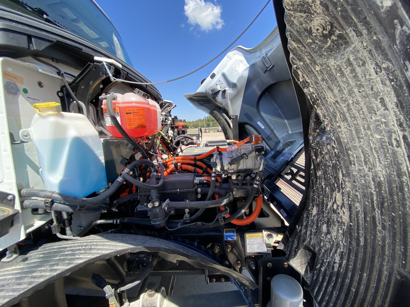 Truck engine bay of a 2024 Mack MD6 E showing coolant reservoir, orange high-voltage cables, and engine components.