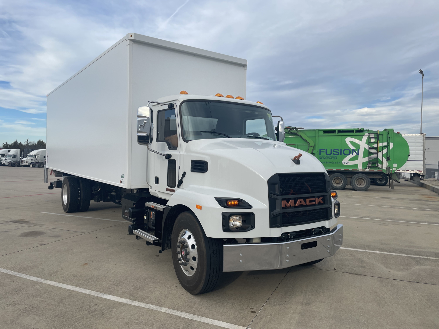 Truck front left view showing a white Mack MD6 E box truck with a large rectangular cargo body, chrome bumper, black grille with MACK letters, orange clearance lights on cab roof, and side mirrors.