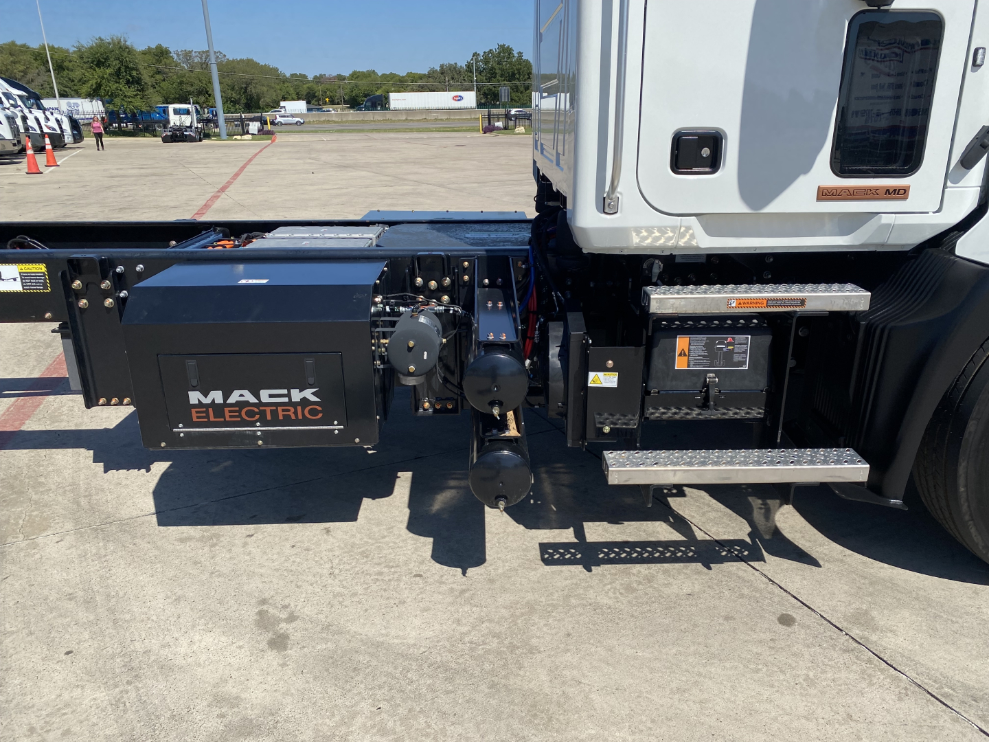 Tractor-trailer chassis and cab of a 2024 Mack MD6 E truck showing Mack Electric battery/auxiliary components under the cab; visible label 'MACK ELECTRIC' on a black box; side steps and wiring near the frame.