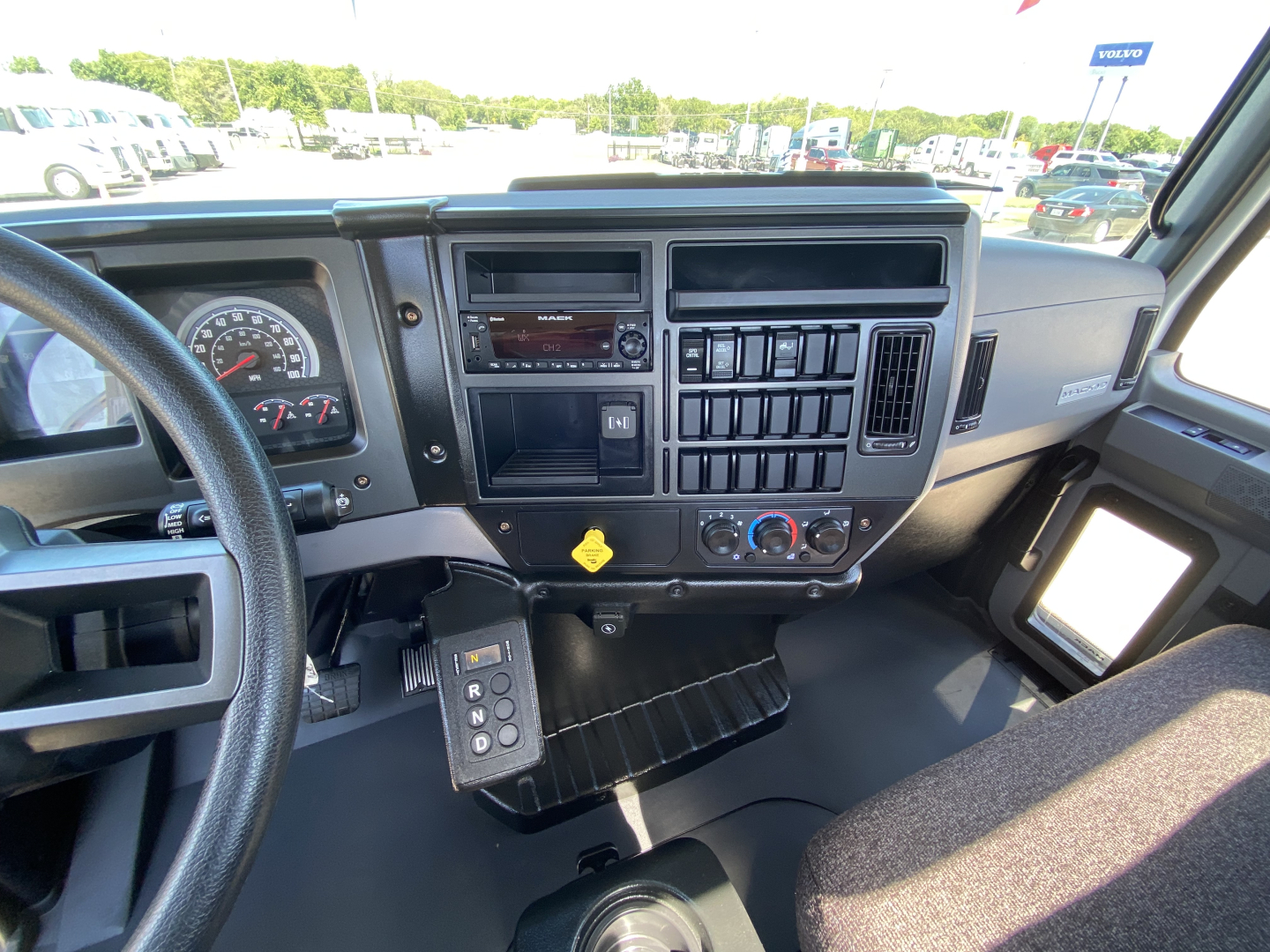 Truck interior dashboard of a 2024 Mack MD6 E showing steering wheel, instrument cluster, radio, HVAC vents, controls, and floor mat.