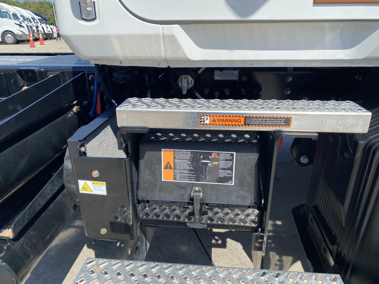 Back of Mack MD6 truck showing storage box with warning label, diamond-plate step, and battery/disconnect box. Visible labels and control box with caution symbols.