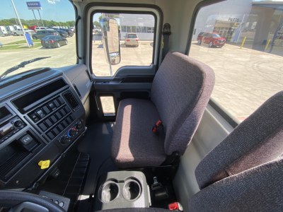 Truck cab interior showing driver and passenger seats, dashboard with control panel and air vents, center console with cup holders, view through windshield to exterior lot.