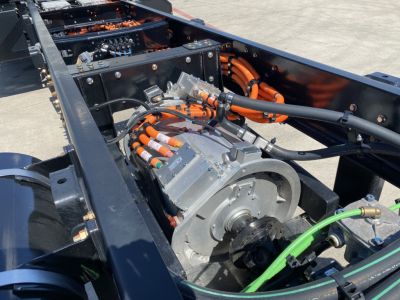 MD6 Mack truck chassis showing electric motor with orange high-voltage cables and green ground strap; battery/drive components visible in open frame.