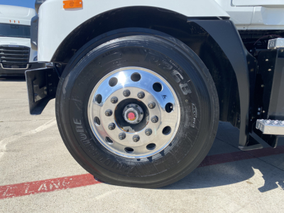 Front wheel and fender of 2024 Mack MD6 E showing chrome wheel with lug nuts and tire, part of truck in service lot.