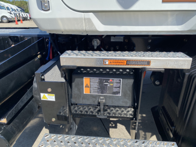 Back of Mack MD6 truck showing storage box with warning label, diamond-plate step, and battery/disconnect box. Visible labels and control box with caution symbols.
