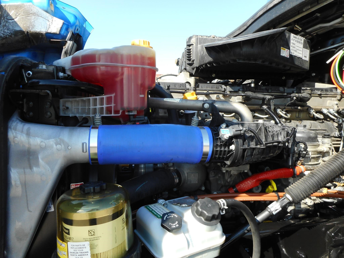 Truck engine bay showing blue air intake hose, coolant reservoir, gold oil/air filter canister, and various hoses and components in a Freightliner Cascadia engine bay.