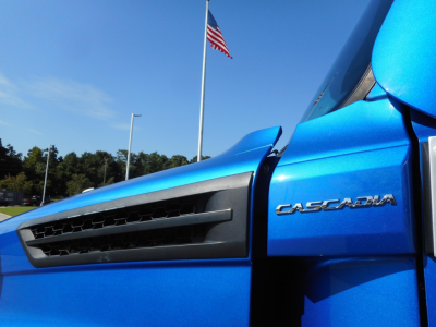 Blue Freightliner Cascadia 126 truck cab side with CASCADIA emblem and front vent, US flag in background.