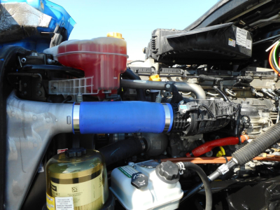 Truck engine bay showing blue air intake hose, coolant reservoir, gold oil/air filter canister, and various hoses and components in a Freightliner Cascadia engine bay.