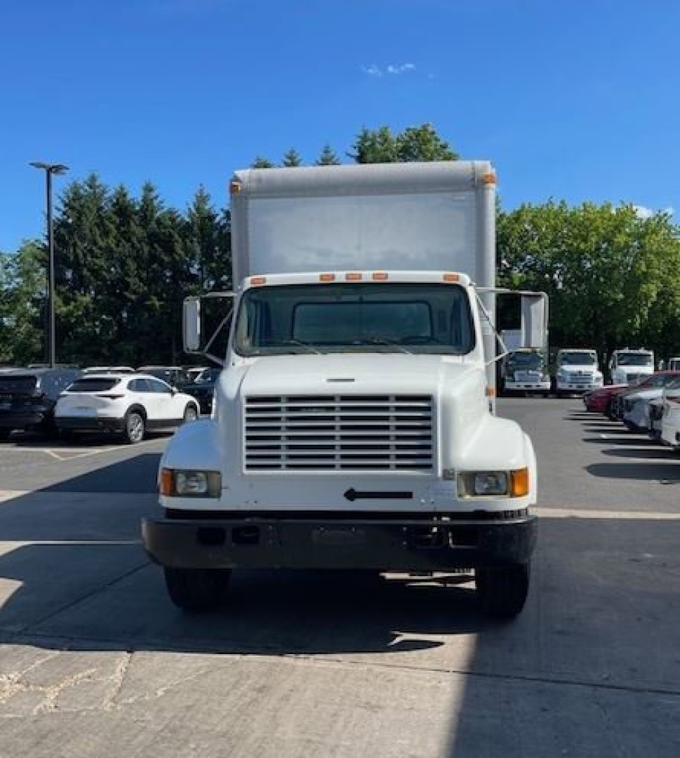 Truck front view showing large rectangular cargo box with grille, headlights, and bumper. White International 4000 series box truck in a parking lot.
