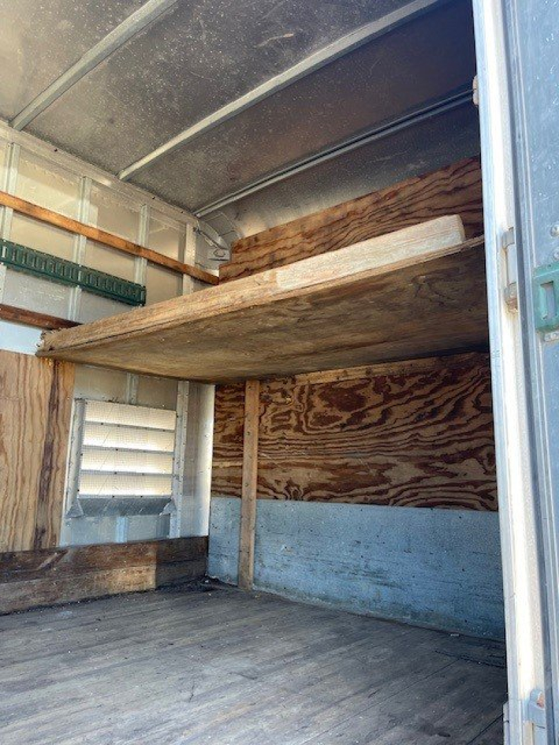 Truck cargo area interior with wooden bulkhead and bunk shelf in a box van; plywood walls, metal framing, green strap rack, wheel well access.
