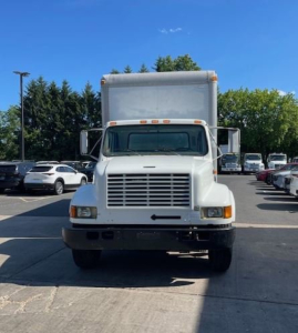 Truck front view showing large rectangular cargo box with grille, headlights, and bumper. White International 4000 series box truck in a parking lot.