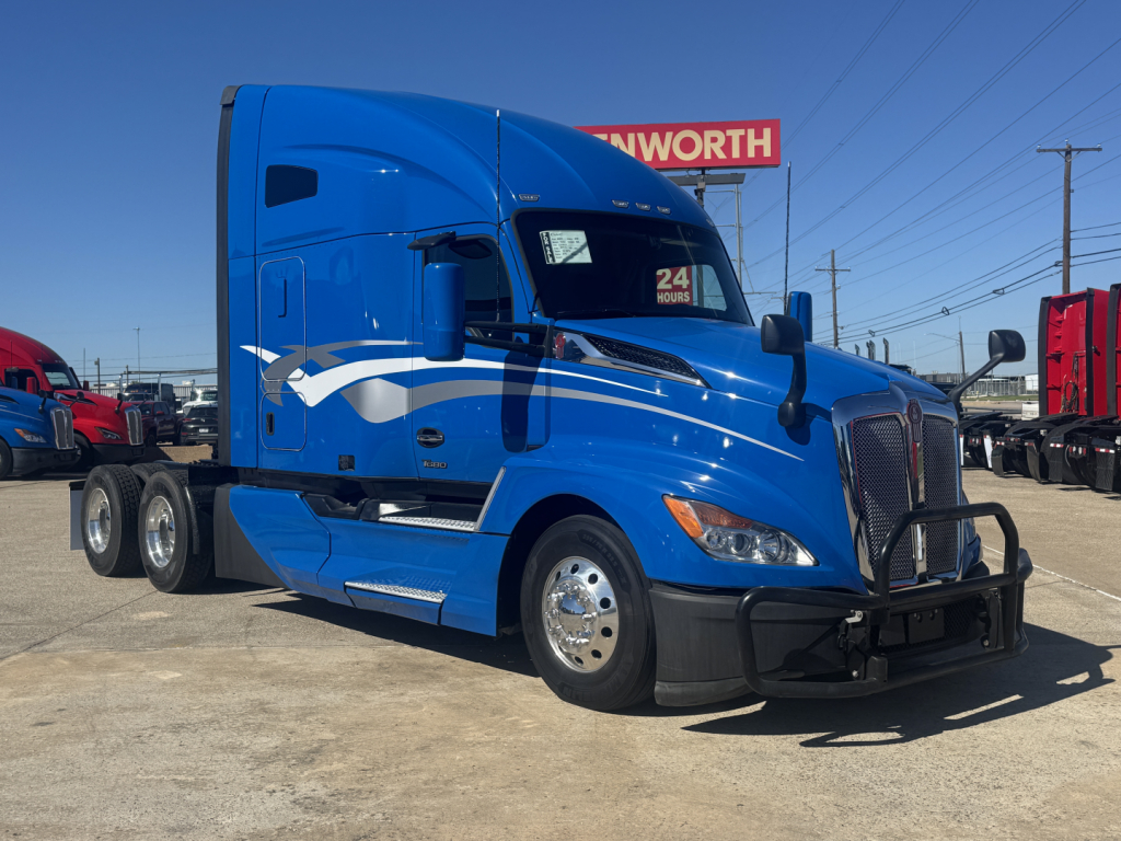 Blue Kenworth T680 semi-truck in a dealership lot, side view, with white and blue graphics, 3 axles behind cab, clear weather.