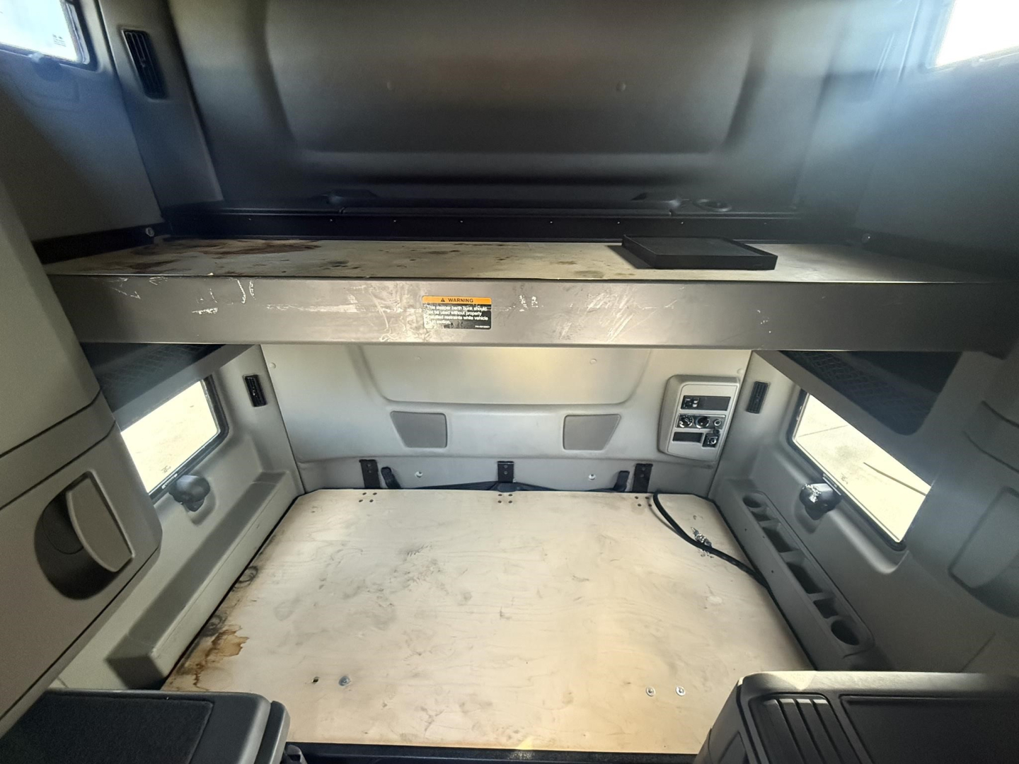 Interior of a used truck sleeper cab showing empty cargo area and overhead bunk area with control panel and window trims.