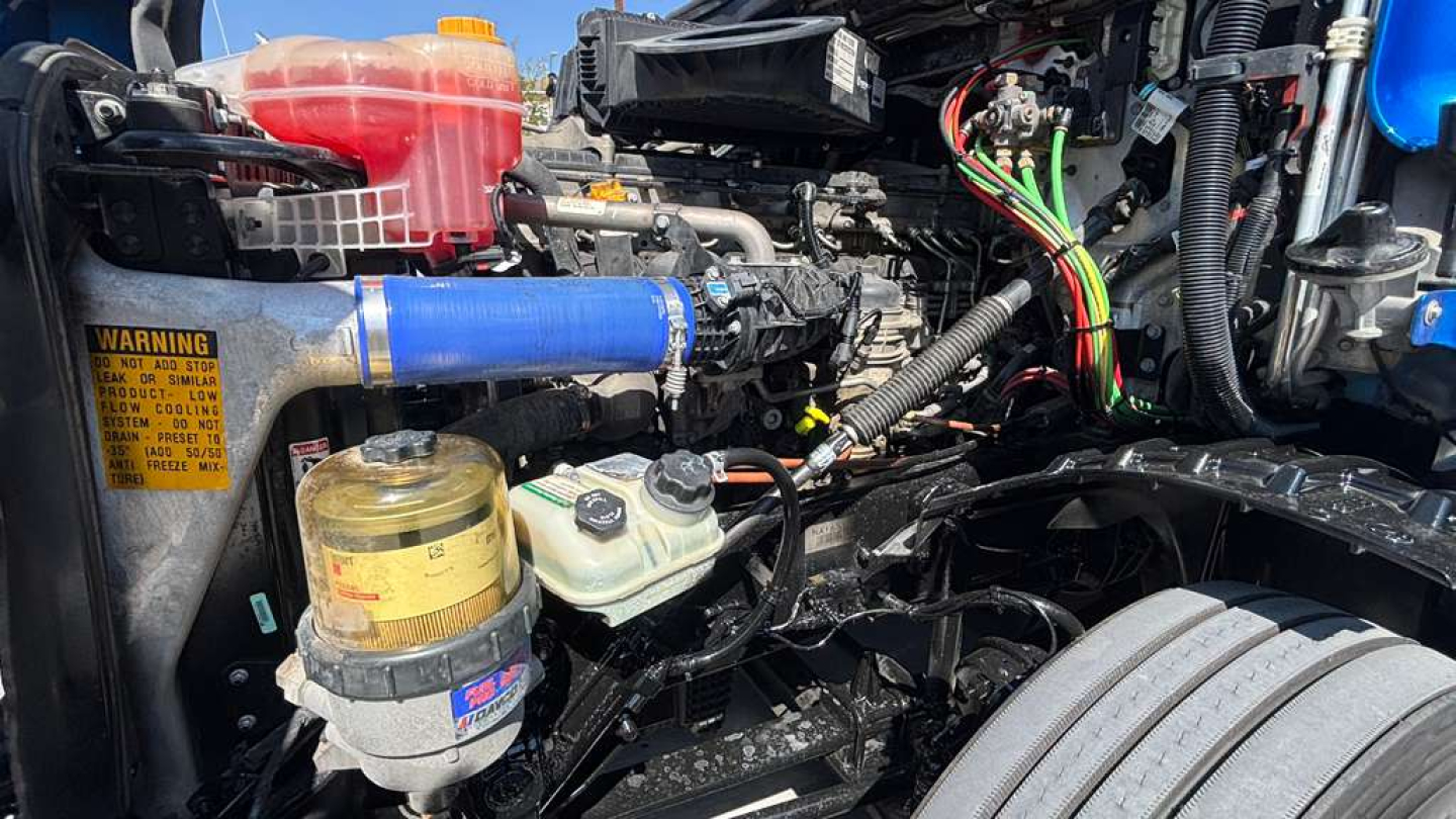 Truck engine bay with various hoses, wiring harnesses, and a blue silicone coolant hose. Visible components include a yellow oil/filters canister, coolant reservoir, power steering fluid reservoir, and a tangle of wiring harnesses.