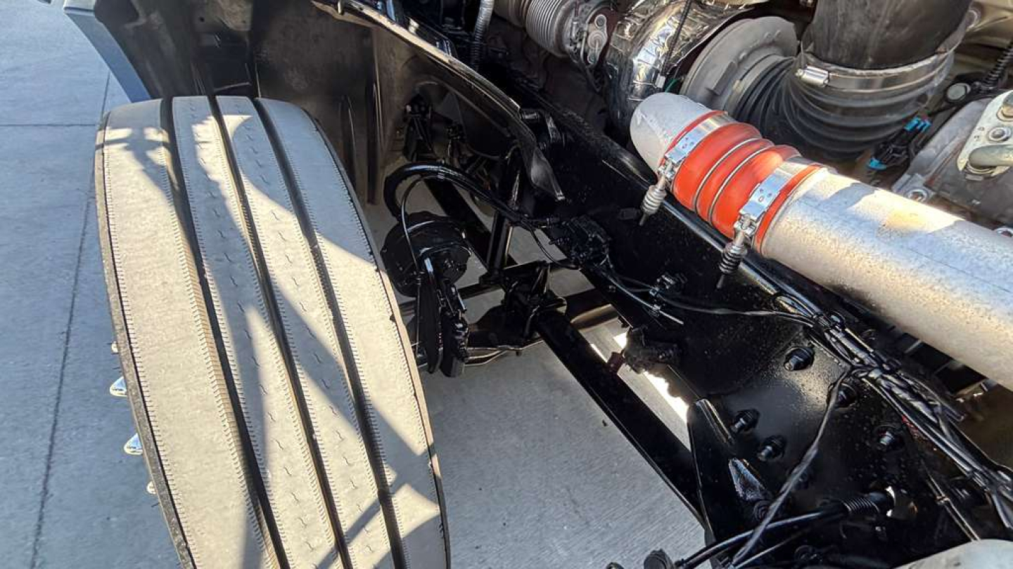 Tractor-trailer engine bay view showing a braided metal hose, chrome fittings, and black chassis components in a Freightliner PT126064ST.
