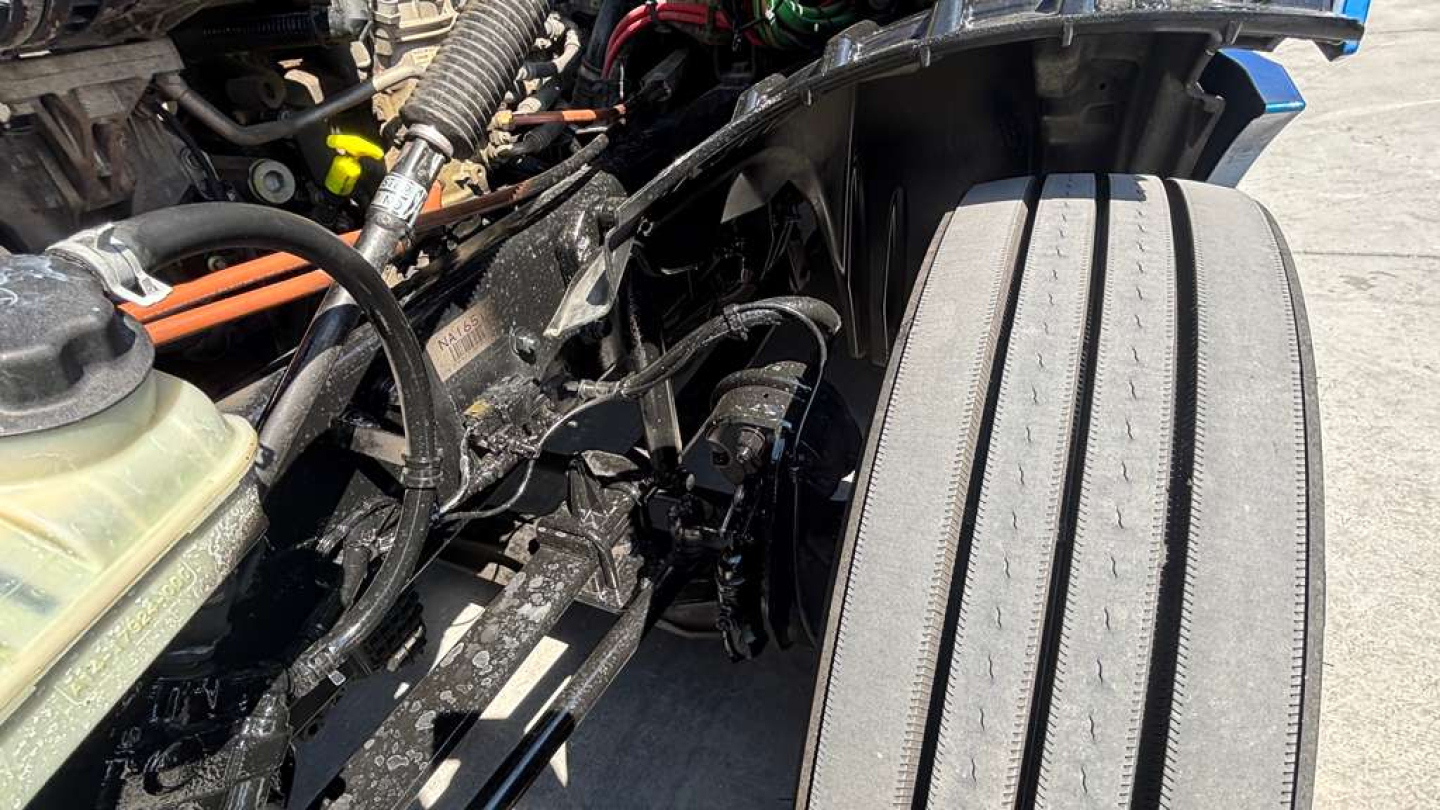 Truck engine bay view with hoses, wiring, and frame; visible air intake hose and coolant reservoir cap; front left wheel well area.