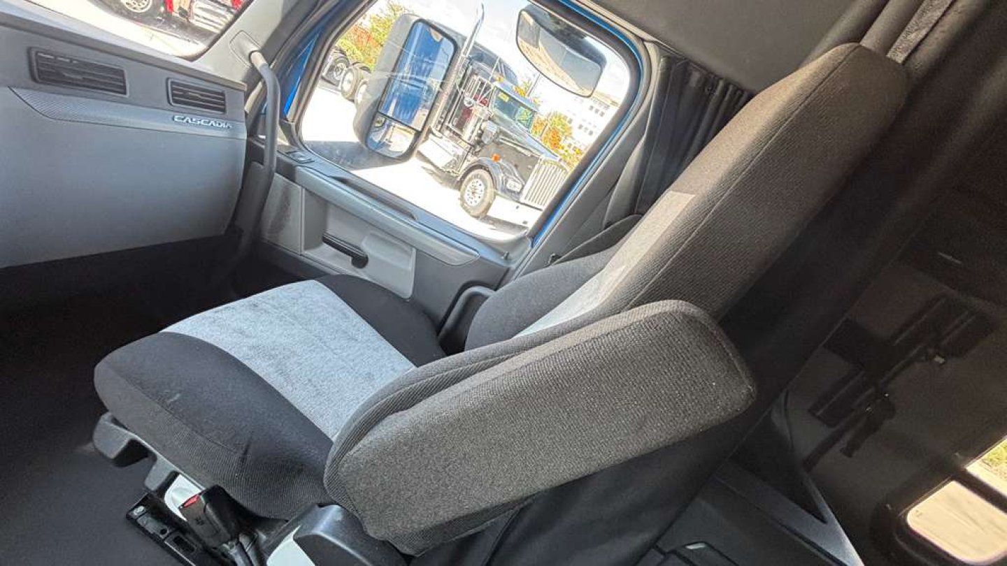 Seat and interior of a Freightliner sleeper truck cab with grey fabric seats and dashboard visible through open door.