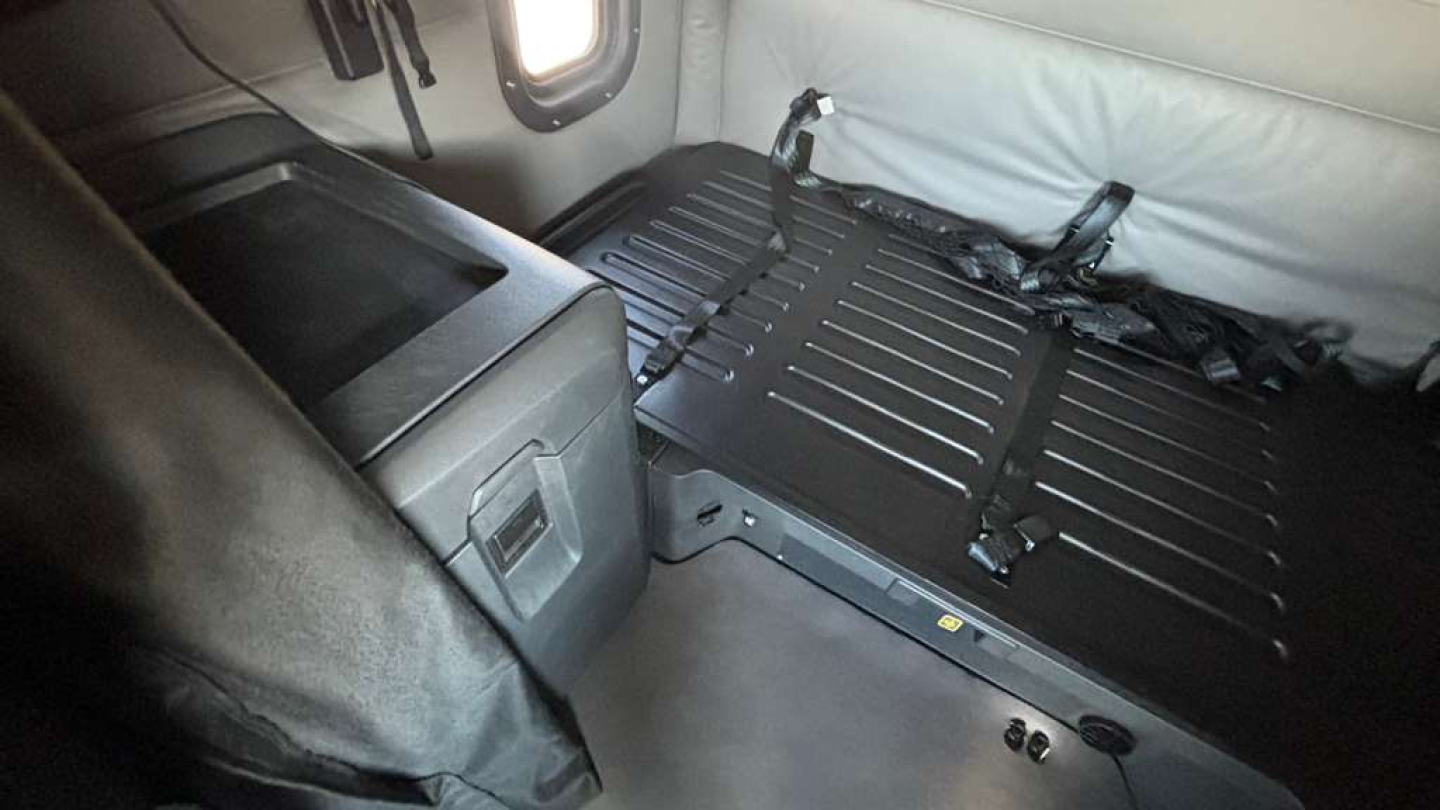 Truck interior showing a folded sleeper bed platform with black straps and surrounding light gray upholstery.